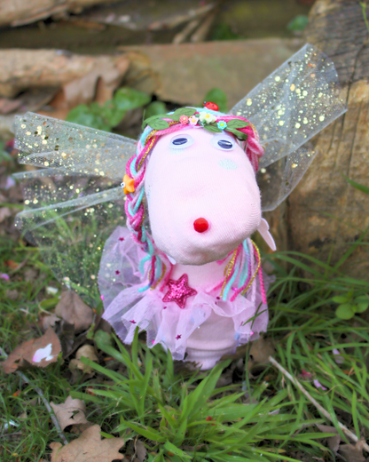 Cheeky the Fairy sock puppet photographed outdoors among greenery, wearing glittery fairy wings and a pink tutu.